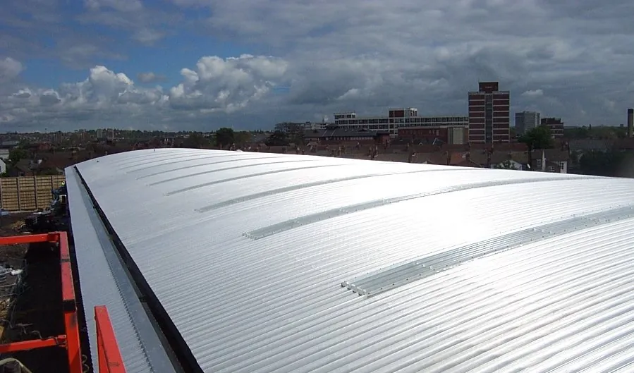 A long curved metal roof with rooflights, seen from the scaffold along its edge, with a town skyline beyond
